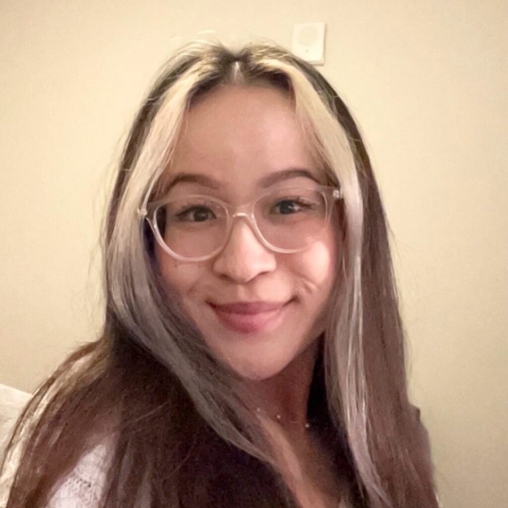 A person with long brown hair and blonde highlights, wearing glasses and a white top, smiles at the camera against a beige background.