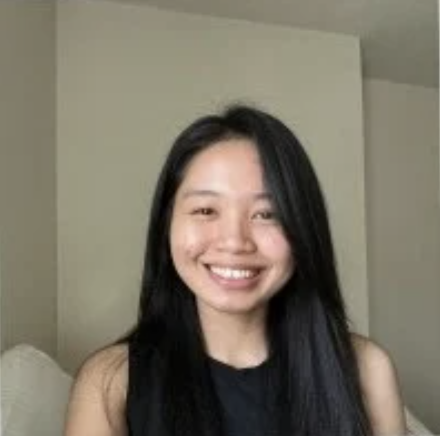 A young woman with long straight black hair smiles at the camera. She is wearing a sleeveless black top and is sitting indoors against a plain light-colored wall.