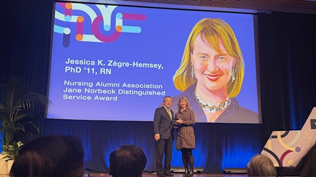 Two people stand on stage smiling as one receives an award. Behind them, a large screen displays a portrait, name, and award title: Jessica K. Zègre-Hemsey, Nursing Alumni Association Jane Norbeck Distinguished Service Award.