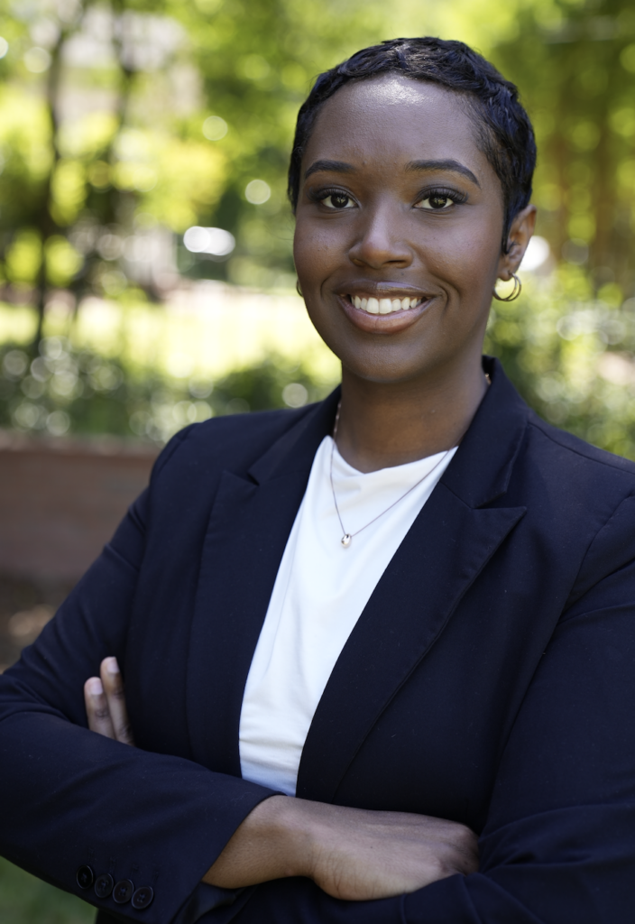 A person with short hair and hoop earrings smiles confidently outdoors, wearing a dark blazer over a white shirt, with arms crossed in front of a blurred green background.