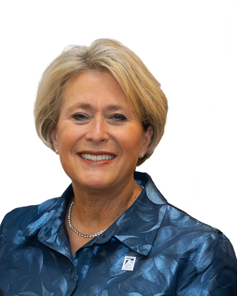A smiling woman with short blonde hair, wearing a blue patterned blouse, a pearl necklace, and a pin on her collar, posed against a white background.