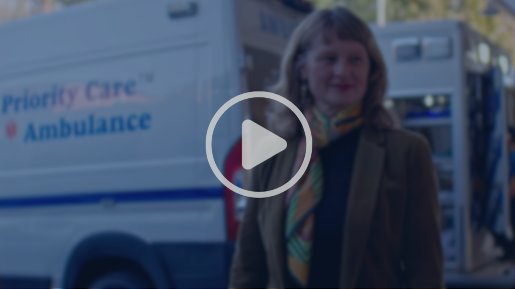 A woman stands in front of an ambulance labeled Priority Care Ambulance, with another emergency vehicle in the background. A play button icon is overlaid on the image.