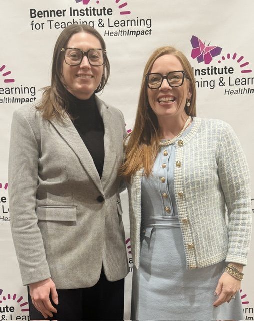 Two women wearing glasses stand and smile in front of a Benner Institute for Teaching & Learning HealthImpact backdrop. One wears a gray blazer and black turtleneck; the other wears a light blue dress with a textured jacket.