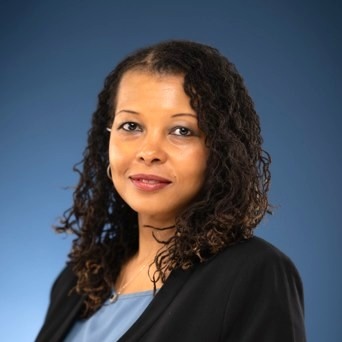 A woman with curly dark hair, wearing a black blazer over a blue top, smiles softly at the camera against a solid blue background.
