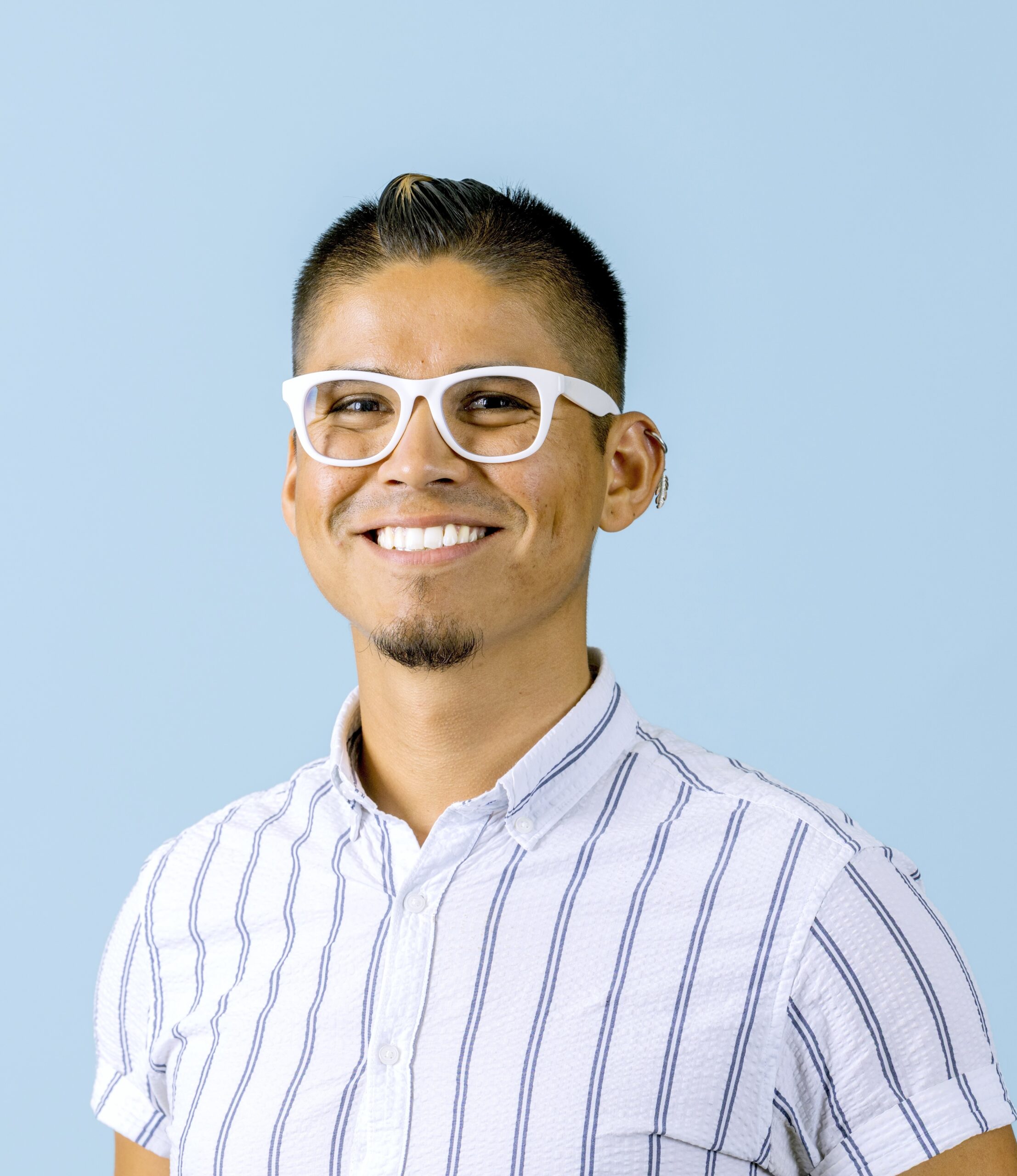 Smiling person with short dark hair, white glasses, and a striped shirt stands in front of a plain light blue background.