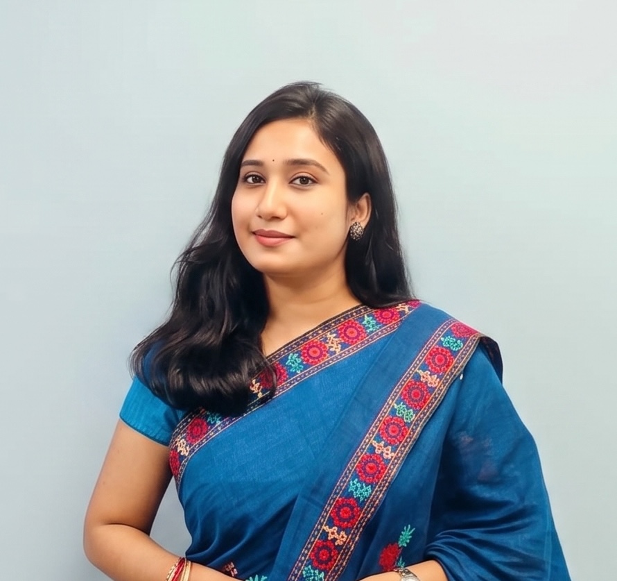 A woman with long dark hair, wearing a blue saree with colorful embroidery, stands against a light blue background, smiling softly at the camera.
