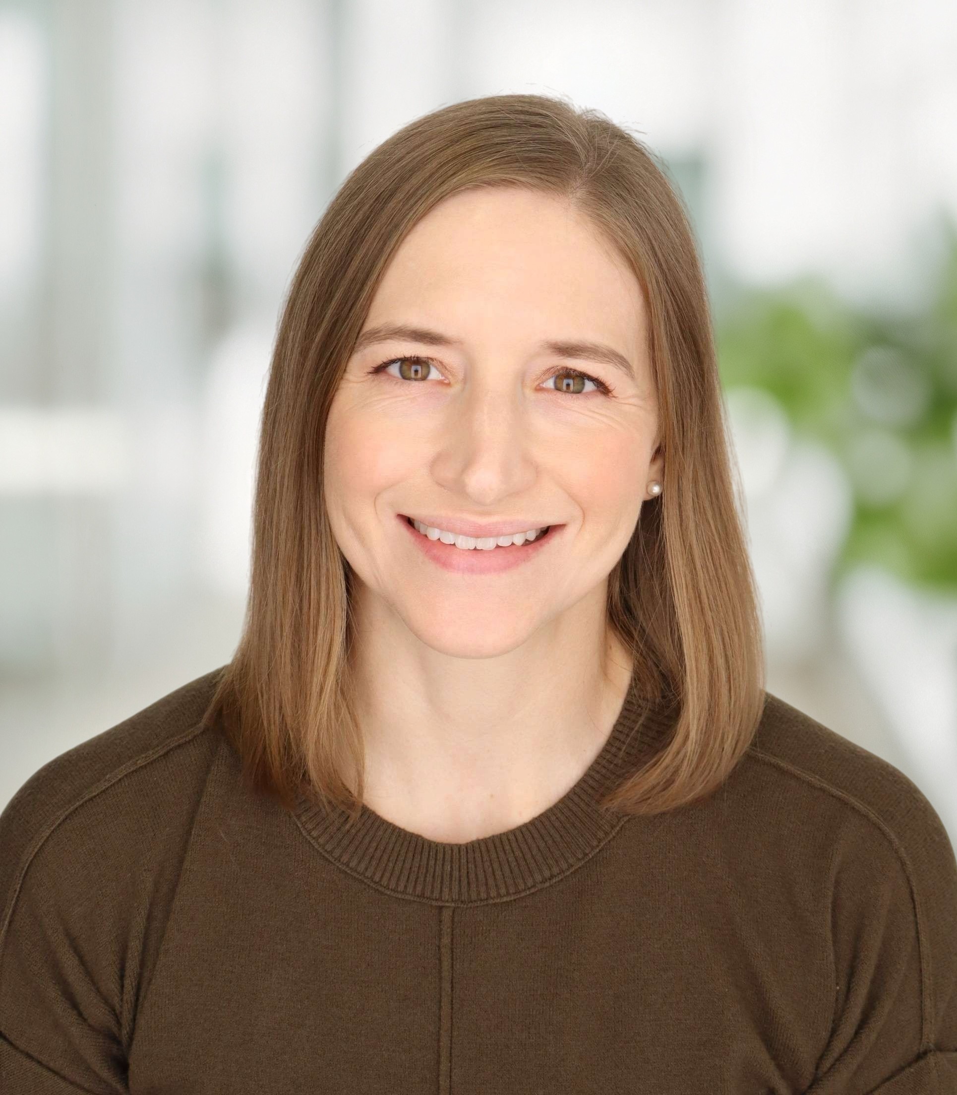 A woman with straight, shoulder-length light brown hair smiles at the camera. She is wearing a dark brown sweater and is in a bright room with blurred green and white background.