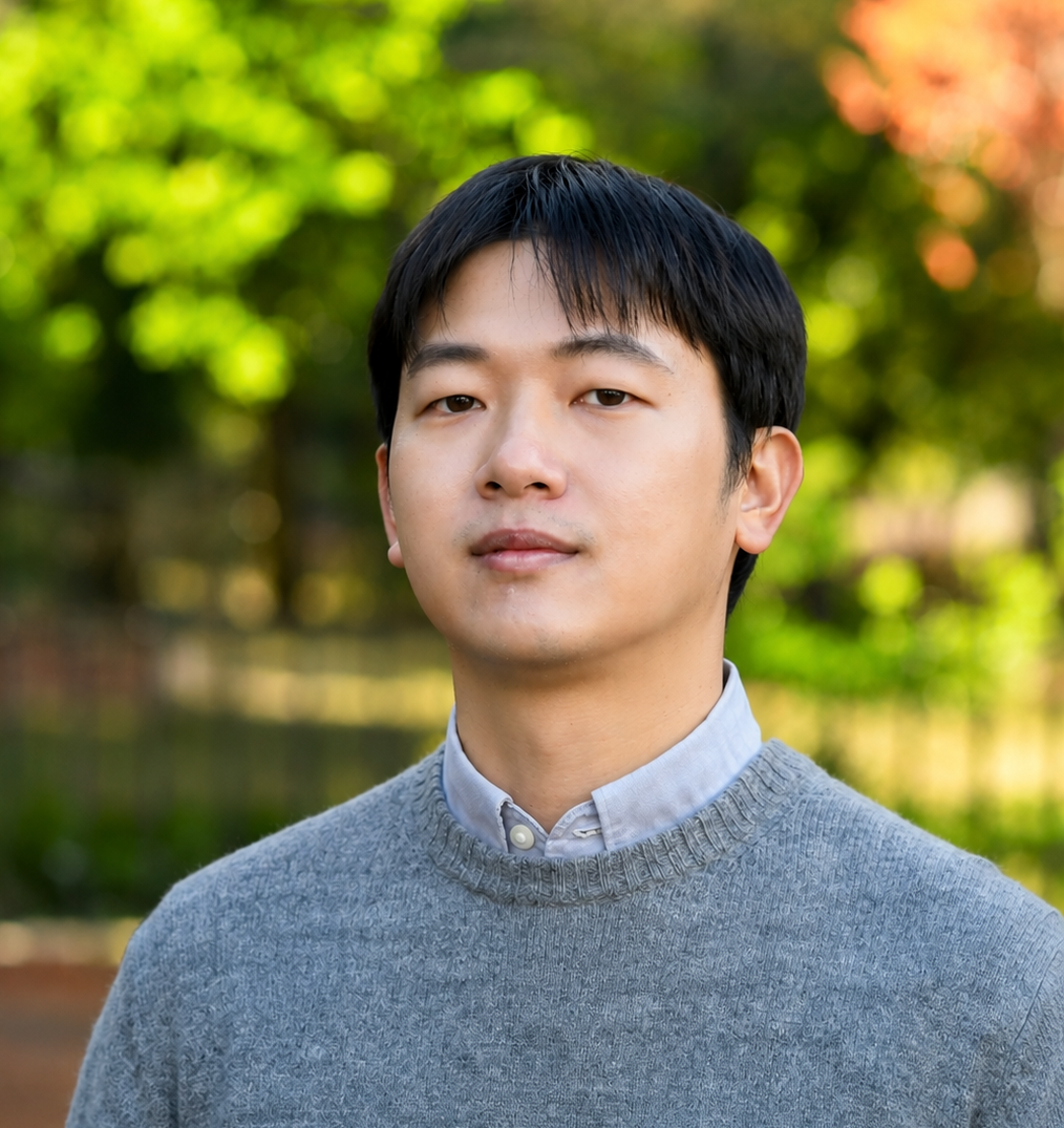 A young man with short black hair wearing a gray sweater over a collared shirt stands outdoors with blurred green trees in the background.