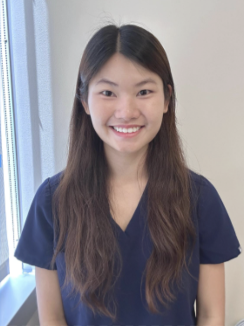 A woman with long brown hair, wearing a navy blue scrub top, stands indoors smiling at the camera. There is a window with blinds to her left.