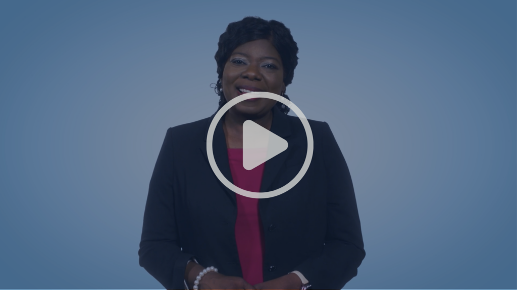 A person wearing a dark blazer and red top stands smiling in front of a blue background, with a large play button icon overlayed in the center, indicating a video.