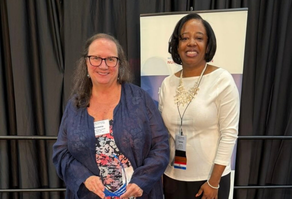 Two women stand indoors, smiling at the camera. The woman on the left holds a glass award. Both wear name badges and business attire, with a banner and black drapes in the background.