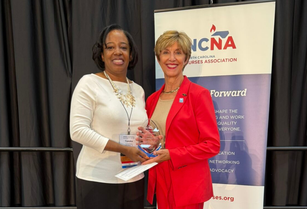 Two women stand smiling; one holds an award. They pose in front of a banner for the North Carolina Nurses Association. Both are dressed professionally, one in a white top and the other in a red suit.