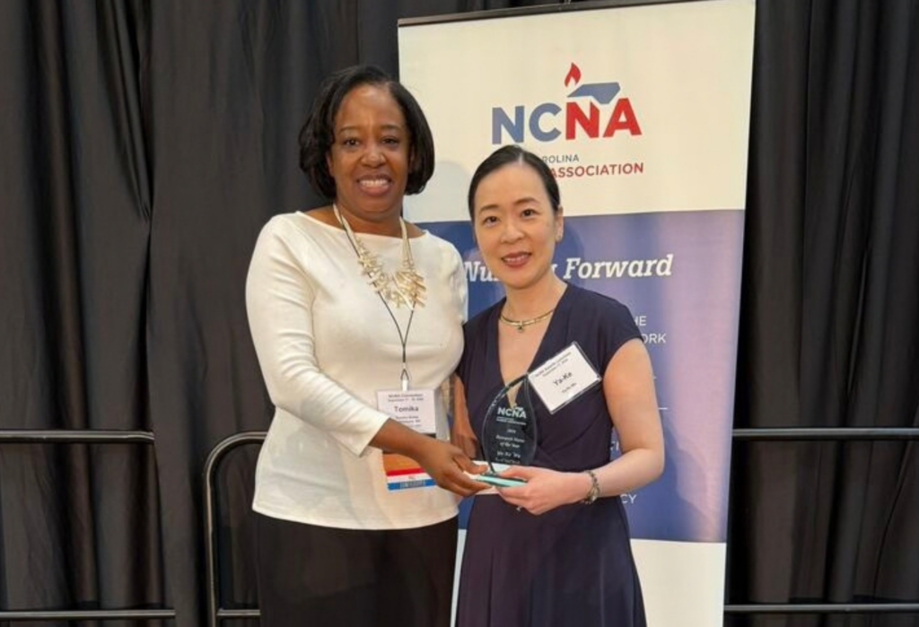 Two women stand in front of an NCNA Association banner. One woman holds an award and both are smiling for the camera at what appears to be a recognition event.