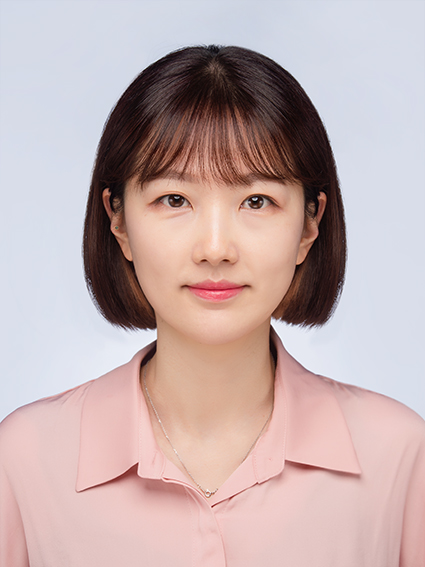 A woman with straight brown hair and bangs, wearing a light pink collared shirt and a delicate necklace, poses against a plain light background.