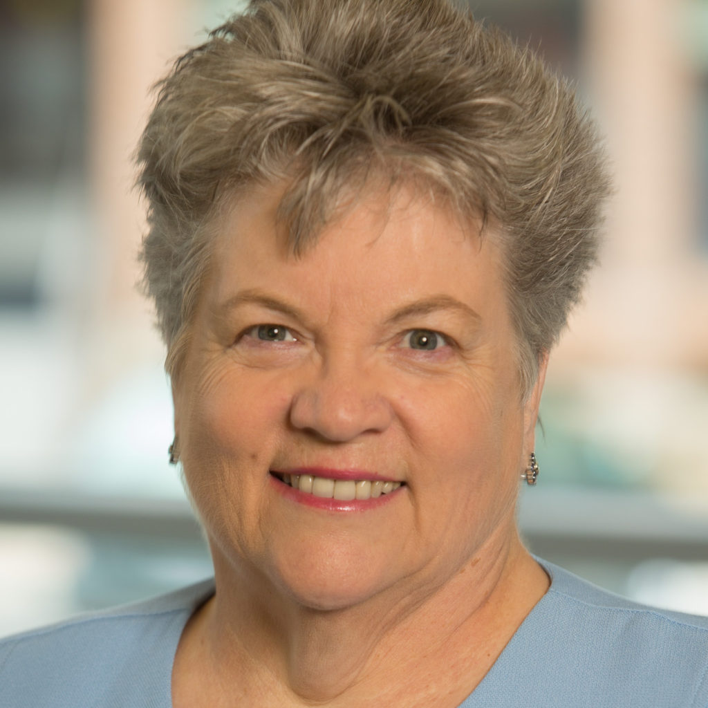 A smiling older woman with short, gray hair wearing a light blue top and small earrings, standing indoors with a blurred background.