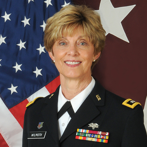 A smiling woman in a U.S. Army dress uniform with medals and badges stands in front of an American flag and a large white star. The name tag on her uniform reads Wilmoth.