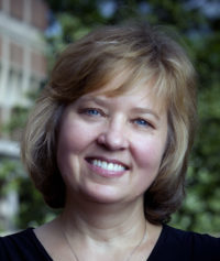 A woman with short, light brown hair and blue eyes is smiling outdoors. She is wearing a black top, and there is greenery and a blurred building in the background.
