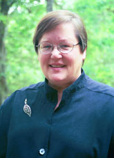 A person with short brown hair and glasses, wearing a dark shirt with a leaf-shaped brooch, stands outdoors in front of greenery and trees, smiling at the camera.